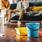 Roof is Leaking, Pipe Rupture at Home: Water Drips into Buckets in Living Room. Angry Couple in Background Calling Insurance Company, Screaming into Phone in Frustration, Trying to Find Plumber
